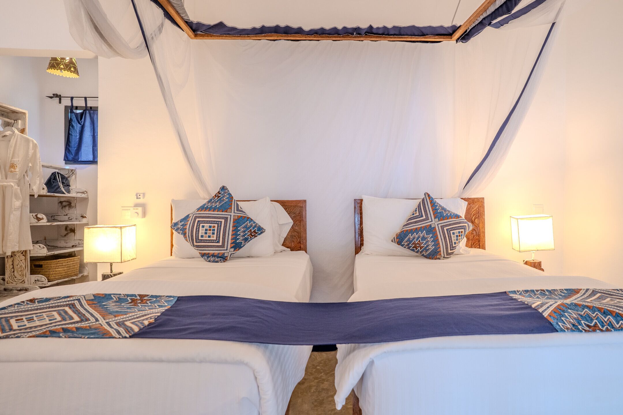 A double room with two beds, each with a patterned pillow, a bedside table with a lamp, and a wardrobe.