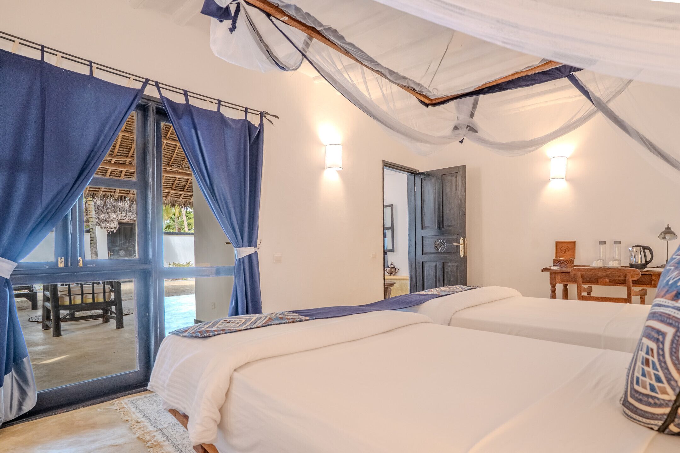Une chambre avec deux lits, une fenêtre avec des rideaux, une porte et une petite table avec une lampe.
