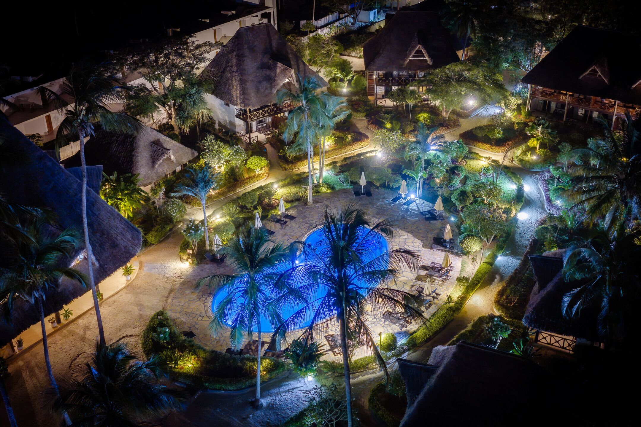 A resort with thatched-roof buildings, a central pool area, and well-lit pathways.