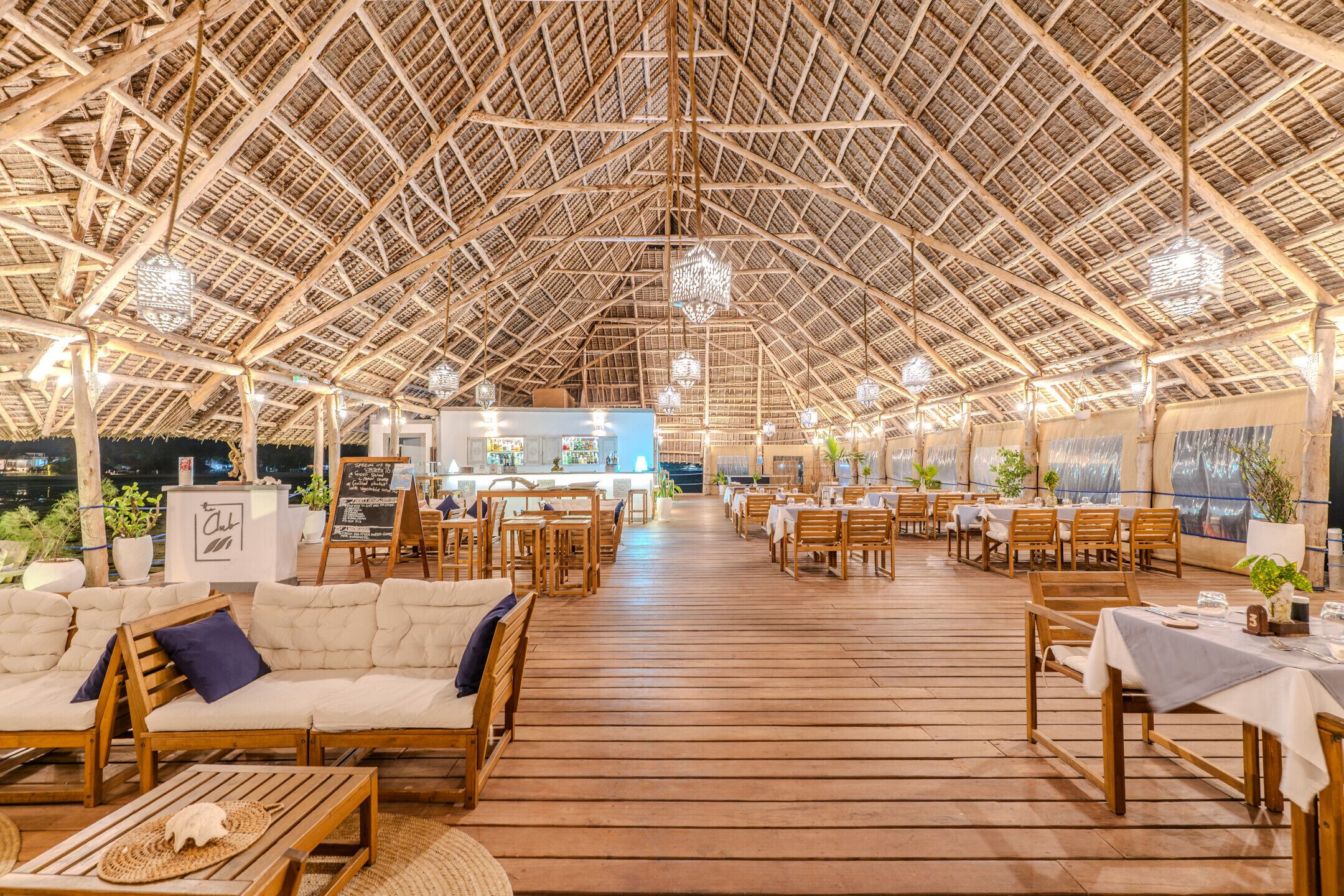 An open-air restaurant with wooden tables and chairs, a bar area, and a thatched roof structure.