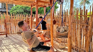 A wooden swing set with ropes and a hanging basket in a sandy outdoor area with a wooden fence and palm trees in the background. - Pongwe Bay Resort (Pongwe)