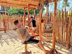 A wooden swing set with ropes and a hanging basket in a sandy outdoor area with a wooden fence and palm trees in the background. - Pongwe Bay Resort (Pongwe)
