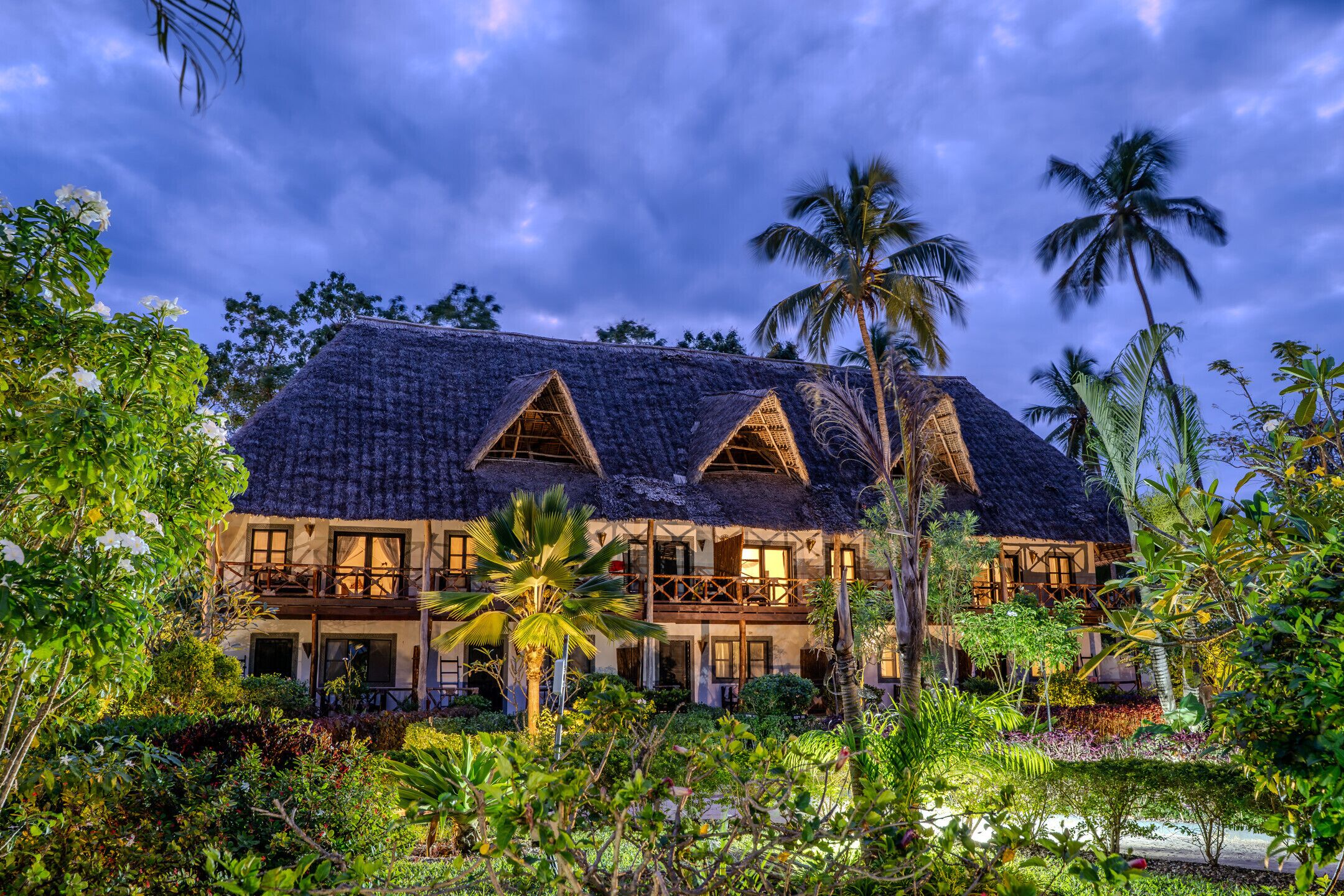 A two-story building with a thatched roof, surrounded by lush greenery and palm trees.