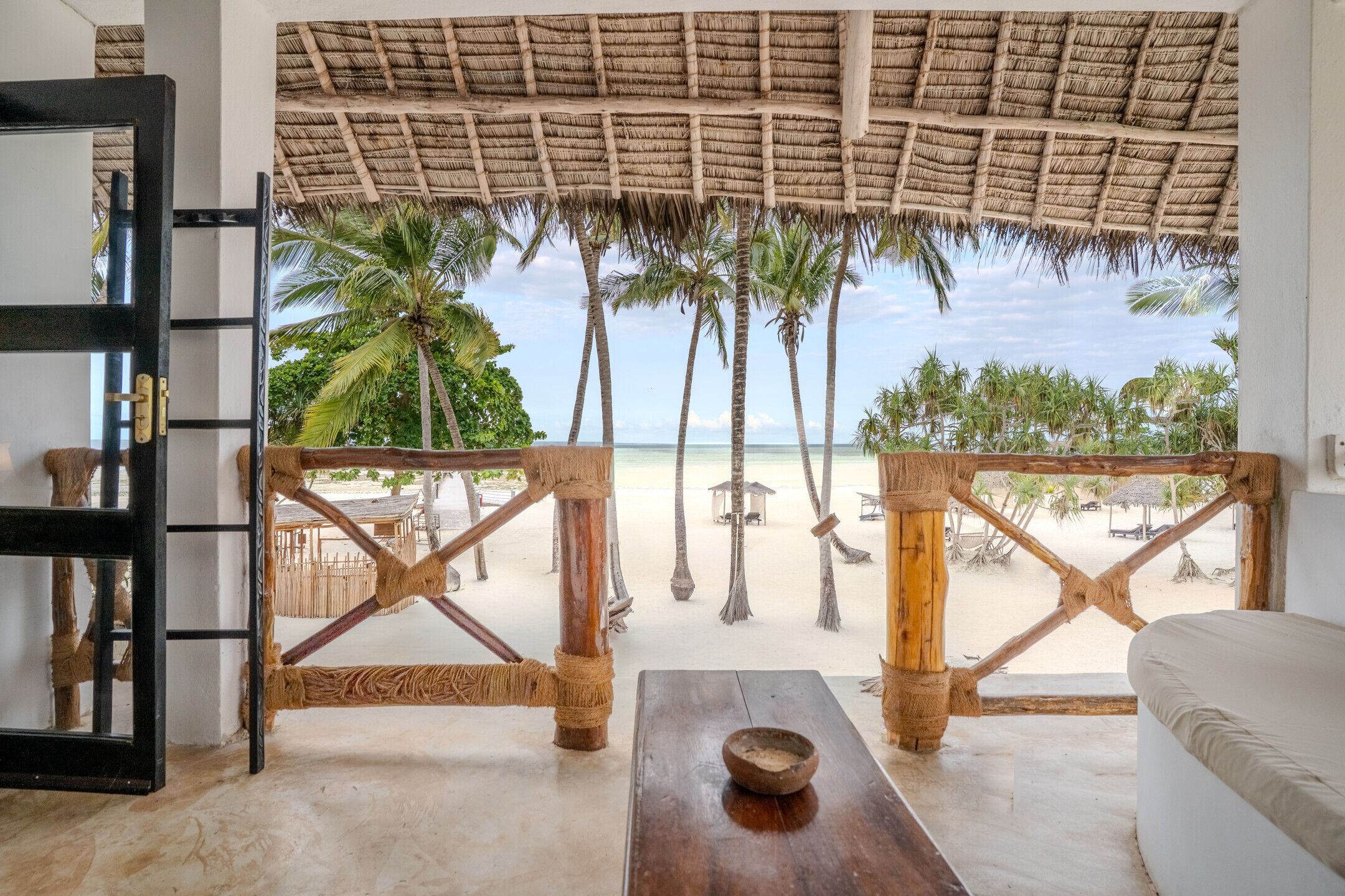Vista a la playa desde una habitación con techo de paja, muebles de madera y vistas a la playa y palmeras.