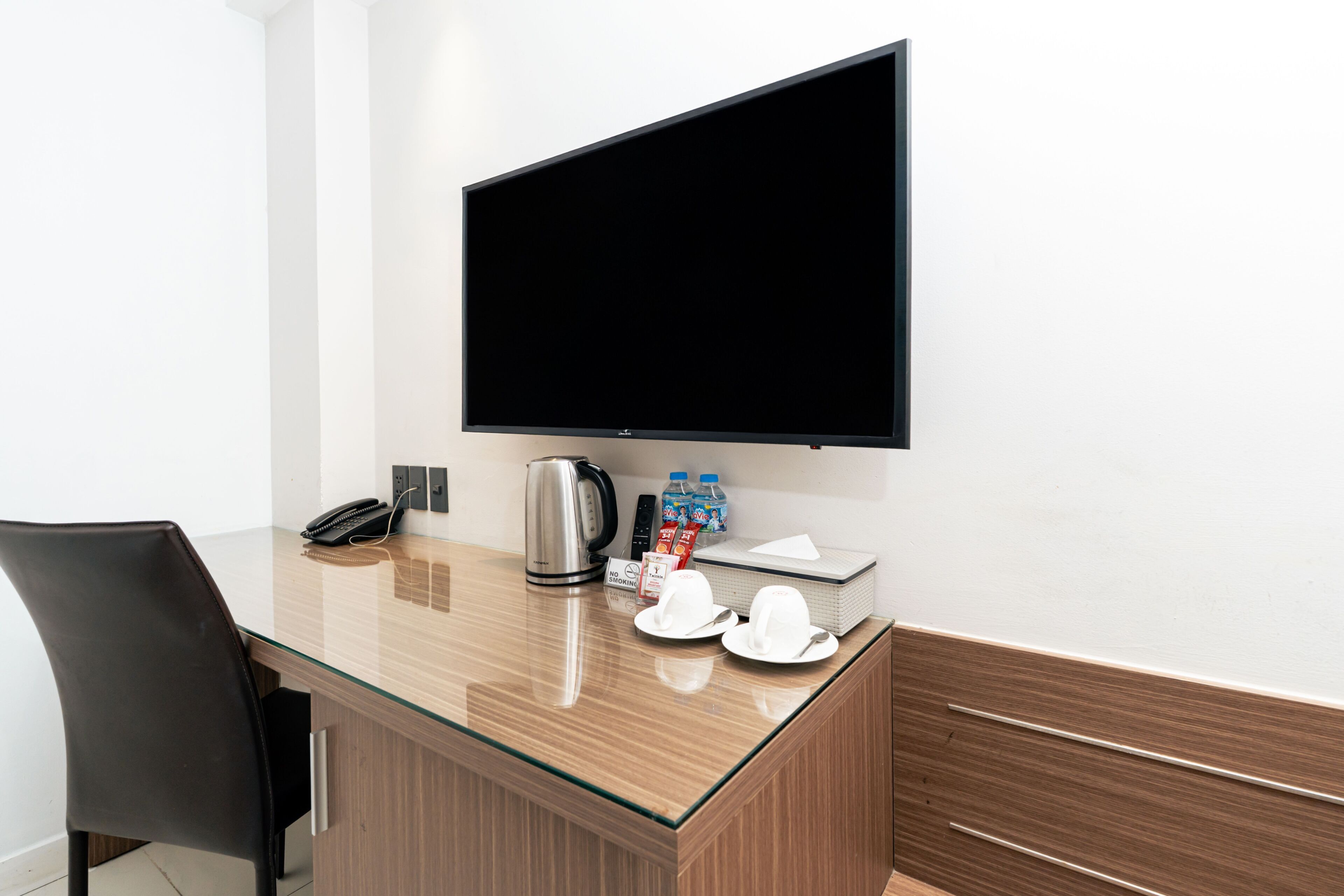 A hotel room with a flat-screen TV mounted on the wall, a minibar with bottled water, a kettle, and a 'No Smoking' sign.