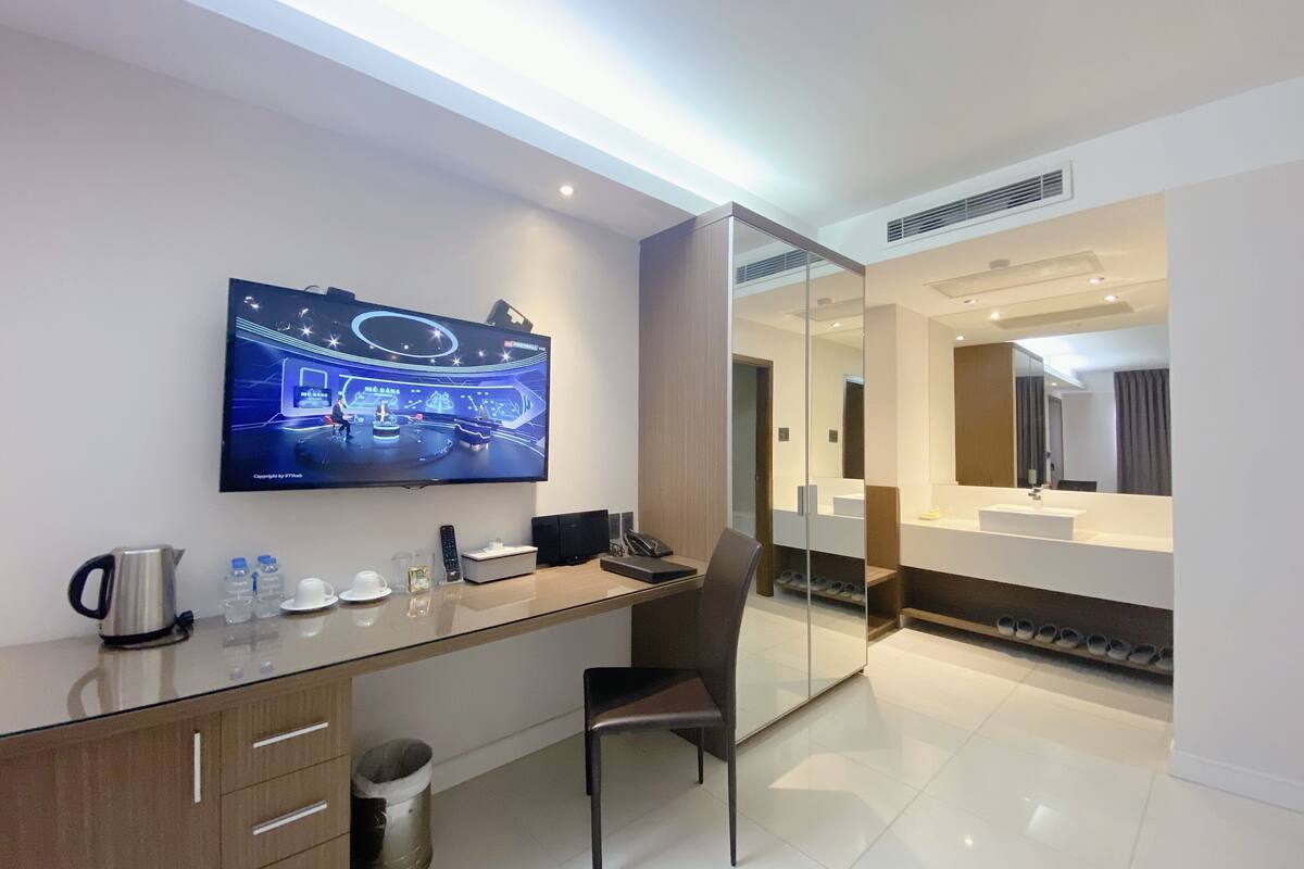 Une chambre d’hôtel moderne équipée d’une télévision à écran plat, d’un bureau avec une chaise, d’un grand miroir et d’une salle de bain séparée.