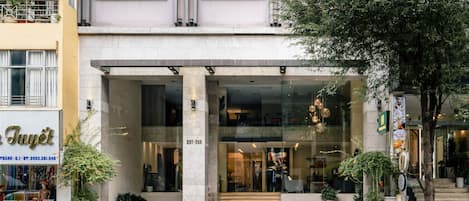 Entrada de hotel con fachada de vidrio, una escalera que conduce a la entrada y un área de descanso al aire libre.