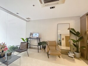 A modern living room with a glass coffee table, a beige armchair, a black desk with a laptop, and a large mirror. There are potted plants and a bookshelf with books. - Nicecy Ben Thanh Hotel (Ho Chi Minh City)