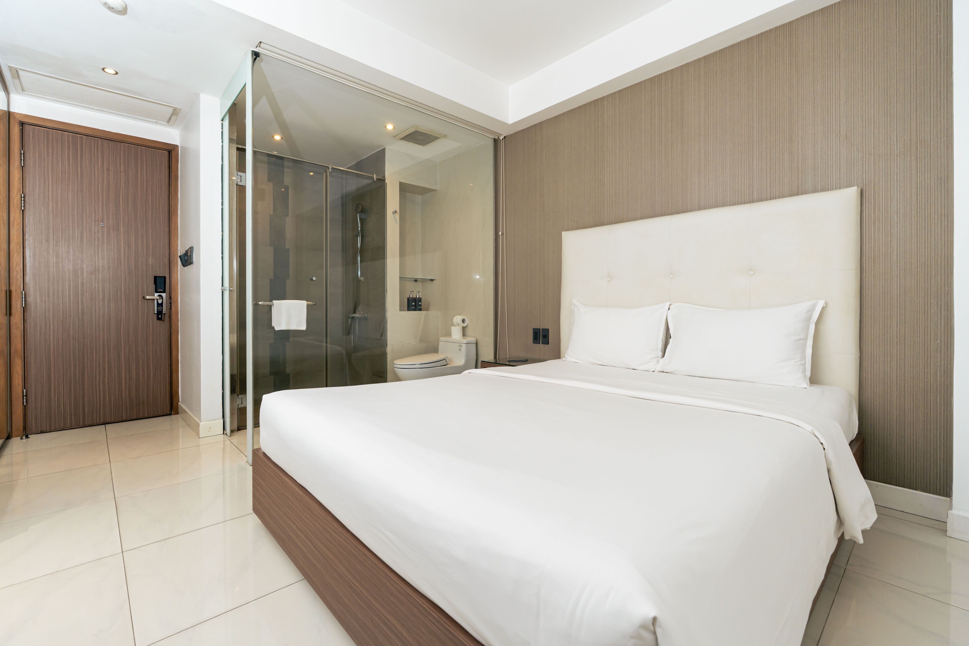 A modern hotel room with a large bed, a glass shower enclosure, a wooden door, and a white tiled floor.