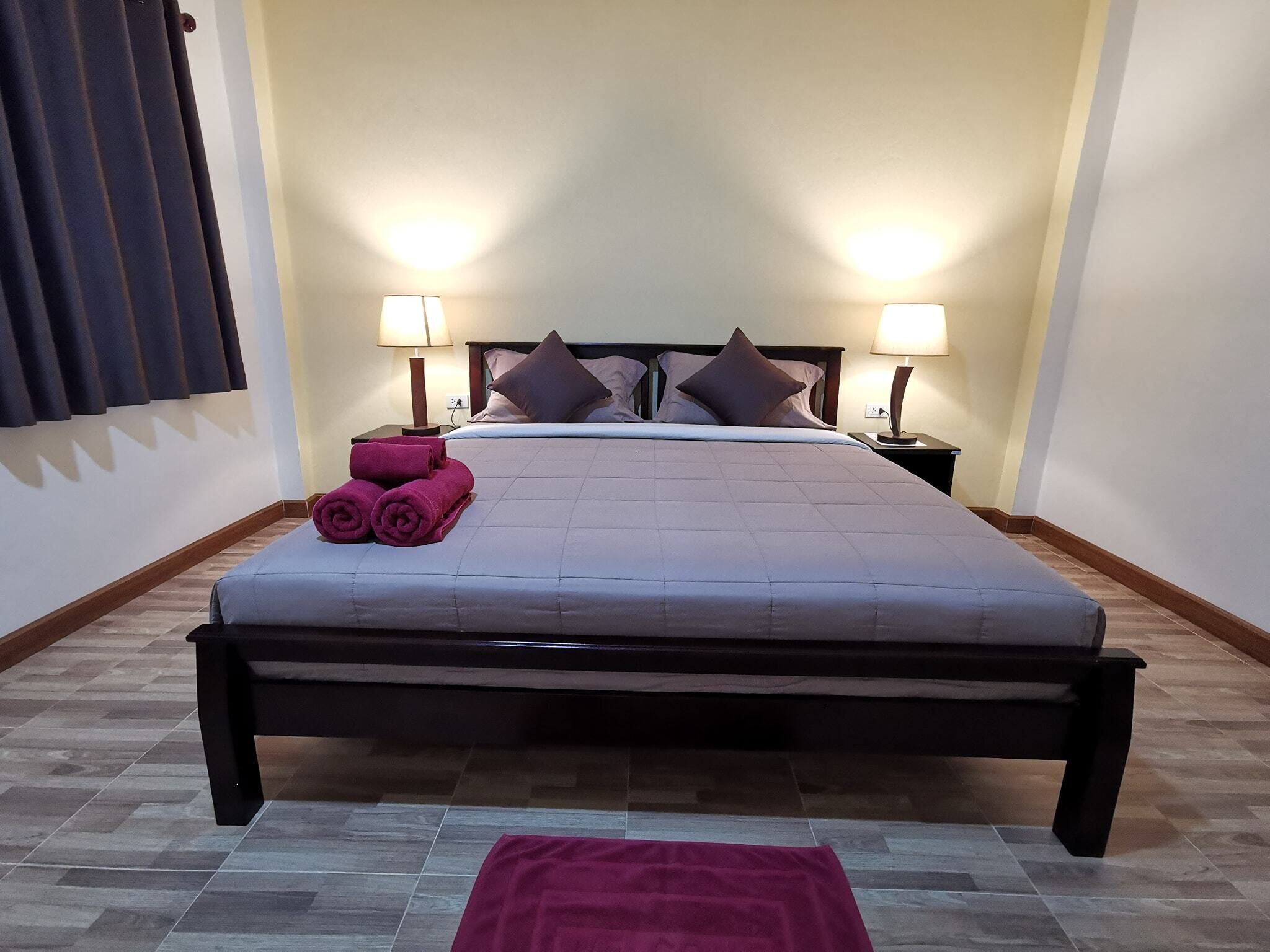 A neatly made bed with a headboard, two bedside lamps, and a pair of rolled towels on the side.