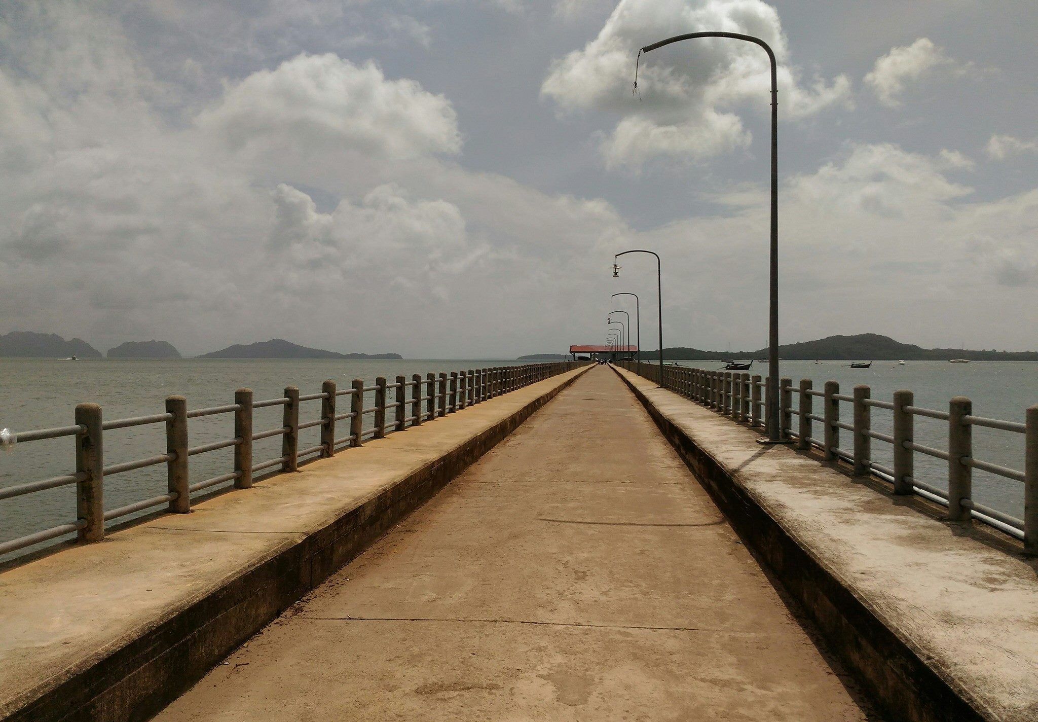 A concrete pier extending into calm water with a metal railing and street lamps.