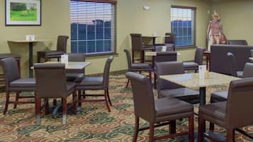 Habitación de hotel con zona de comedor que incluye mesas y sillas, alfombra estampada y un gran ventanal con persianas.