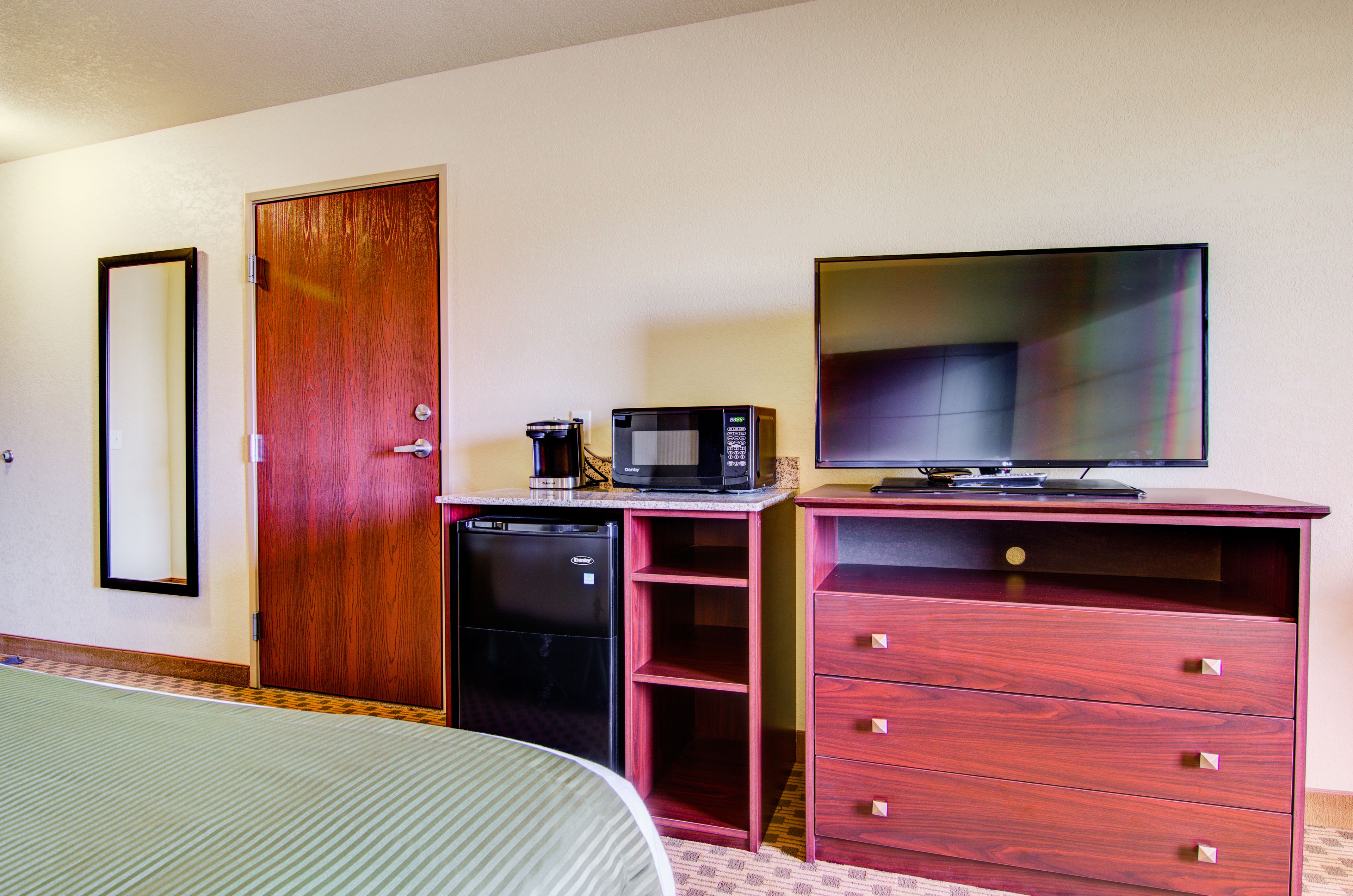 A hotel room with a bed, a wooden door, a kitchenette with a microwave, a coffee maker, and a flat-screen TV on a wooden cabinet.
