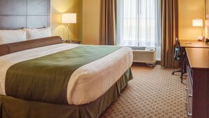 A hotel room with a large bed, a desk with a chair, a lamp, and a window with curtains. - Cobblestone Inn & Suites - Winterset, IA (Winterset)