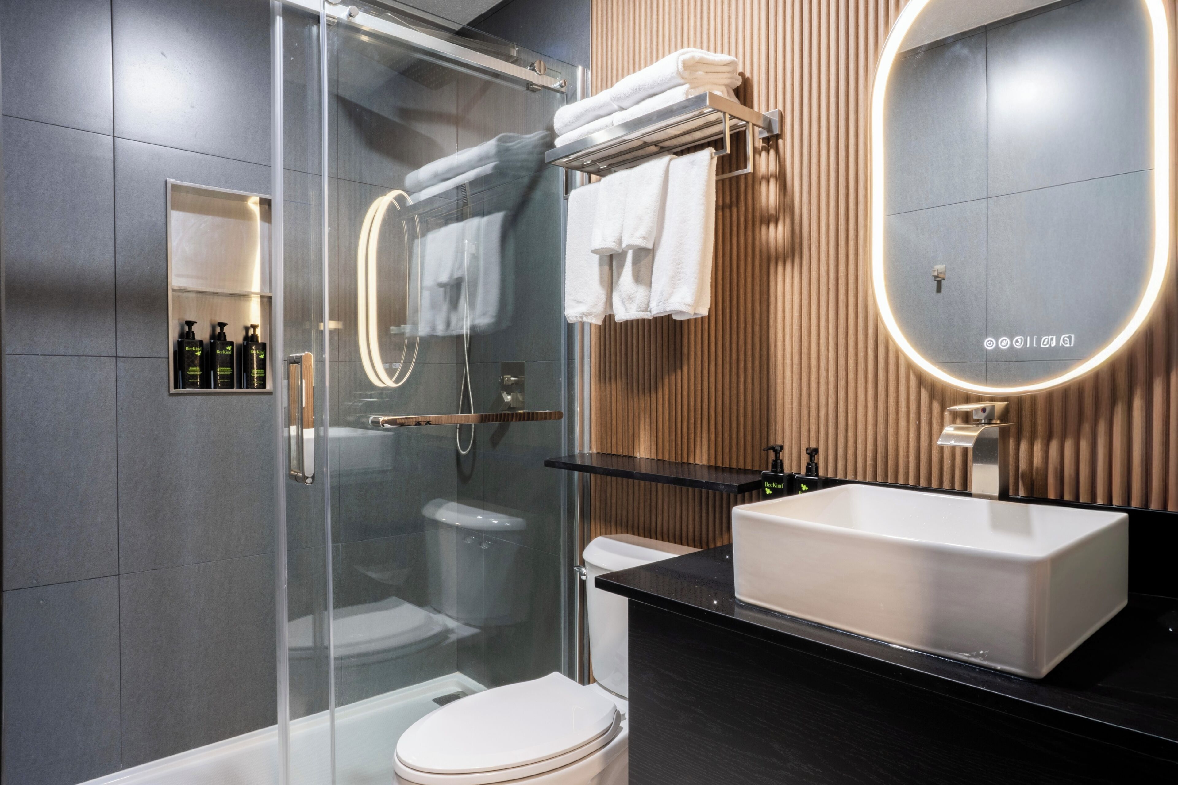 Une salle de bain moderne avec une douche à l’italienne en verre, des toilettes blanches, un comptoir noir, un grand miroir et un panneau mural en bois.