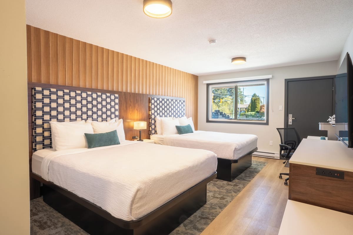 A hotel room with two beds, a desk, and a window with a view of trees.