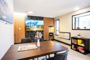 A modern dining area with a wooden table, chairs, a vase with flowers, a newspaper, a wall-mounted TV, a popcorn machine, and a bookshelf with books and games.