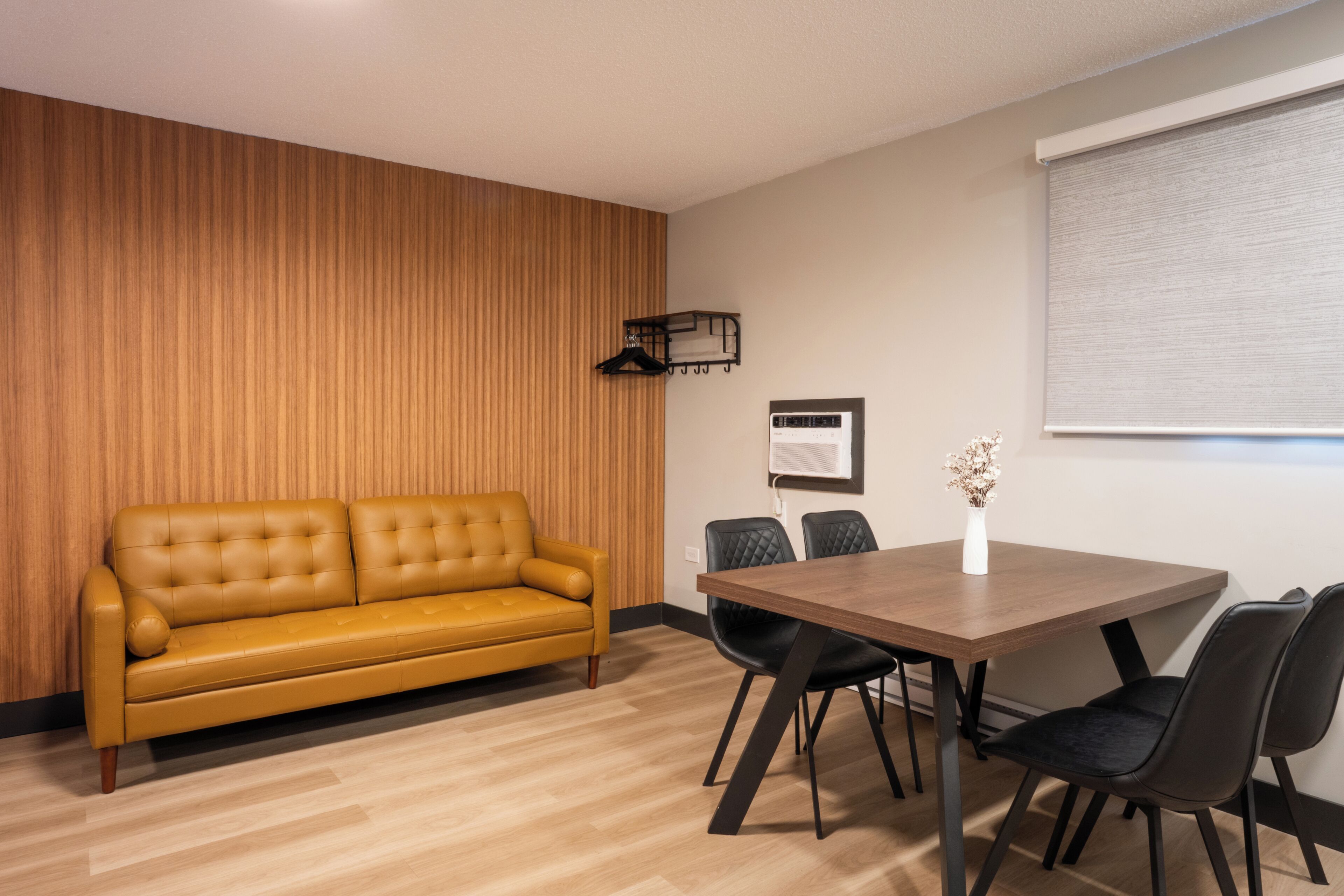 Una habitación moderna con un panel de madera en la pared, un sofá amarillo, una mesa de comedor de madera con cuatro sillas y una ventana con persianas.