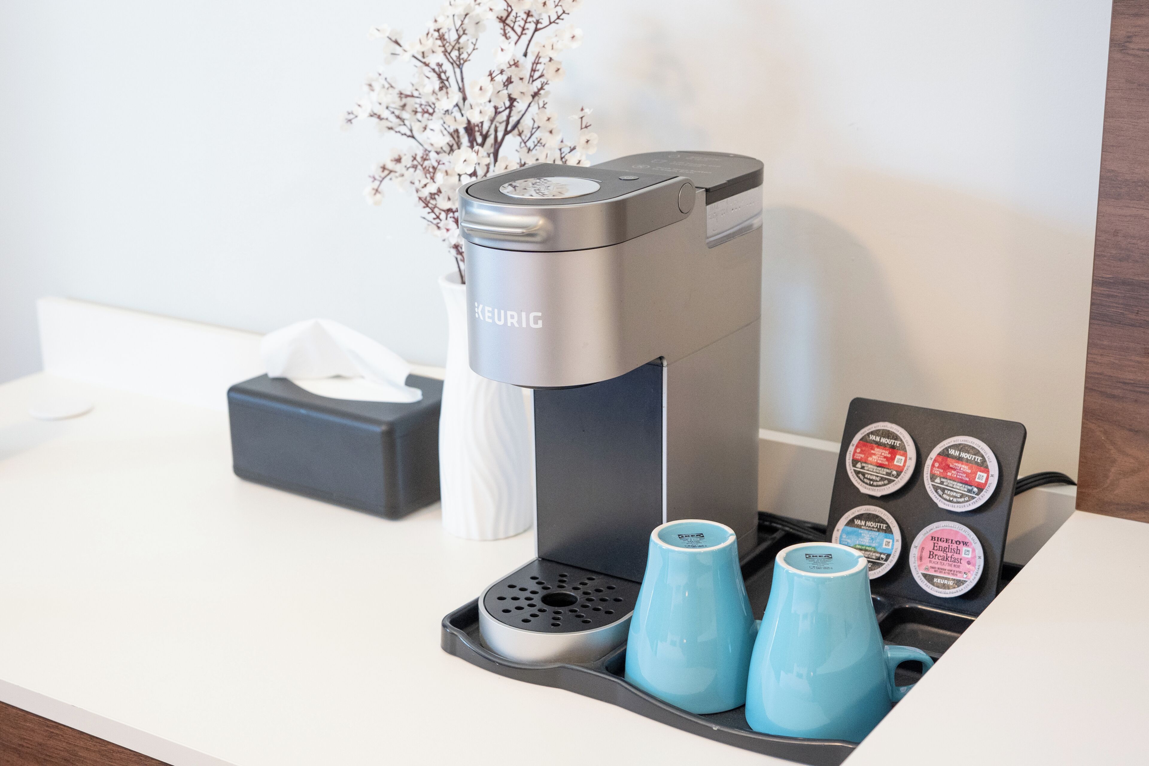 En Keurig kaffemaskine med en holder til kaffepuder, en tissueske og to blå kopper på en hvid bordplade.