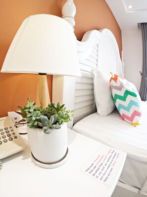 Une chambre d’hôtel bien rangée, avec un lit, une lampe de chevet, une plante en pot, une télécommande et un mot posé sur la table.