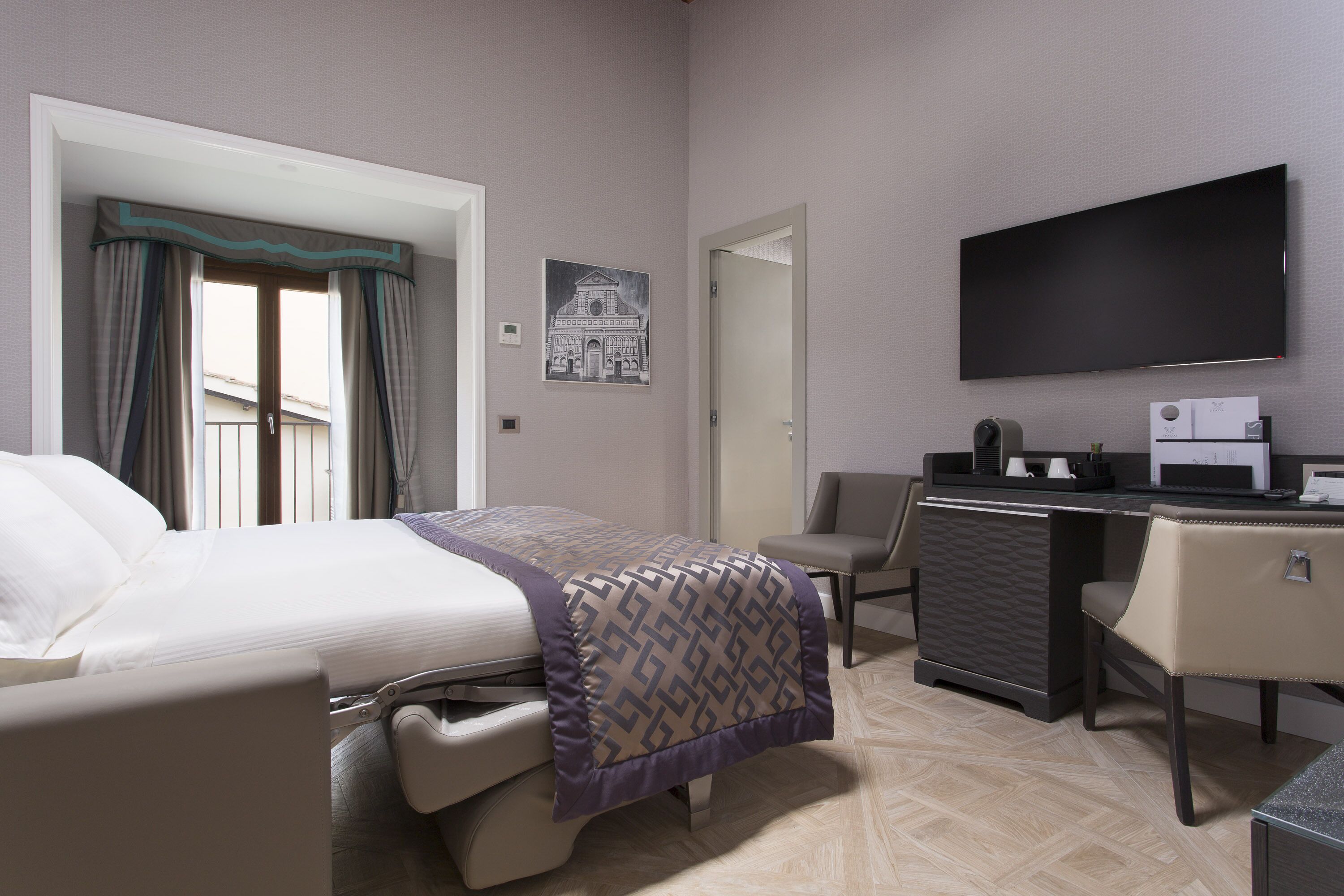 A hotel room with a bed, a desk, a chair, a flat-screen TV, and a painting on the wall.