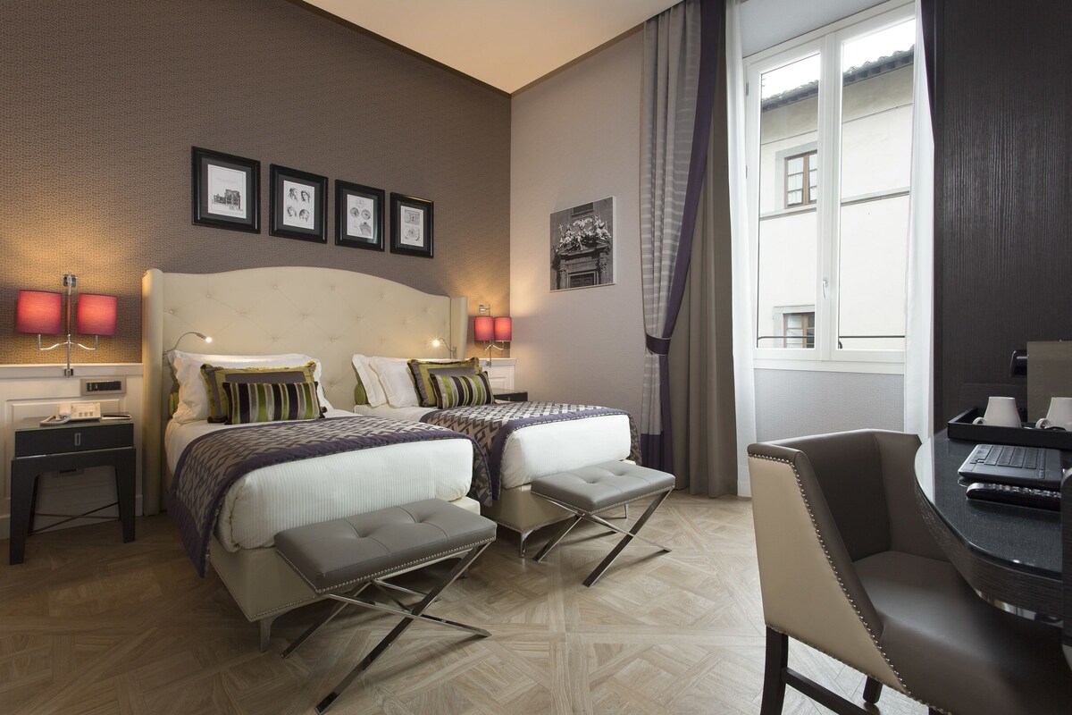 A hotel room with two beds, a desk, and a chair. There are framed pictures on the wall and a window with curtains.