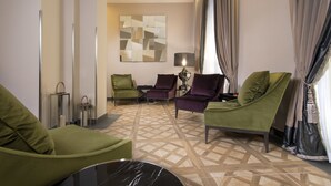 A modern hotel room with a geometric patterned floor, green and purple armchairs, a black marble table, and a large abstract artwork on the wall.