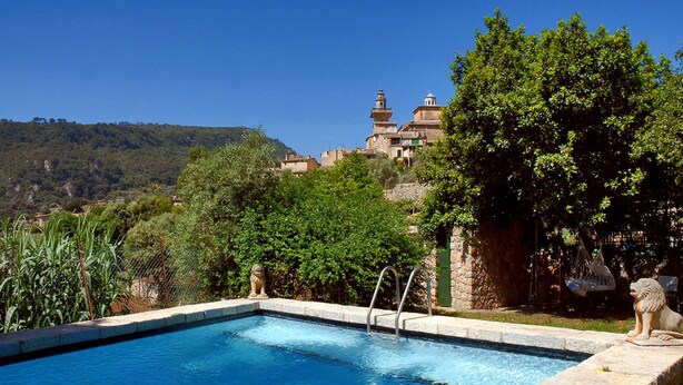 A swimming pool with a stone border, surrounded by lush greenery and a view of a hillside with buildings. -  Sa Vall Turismo de Interior (Valldemossa)
