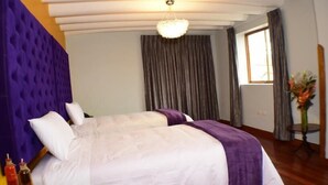 A hotel room with a large bed, a purple headboard, a chandelier, a window with curtains, and a vase with flowers. - Tocapu Boutique Hotel (Cusco)