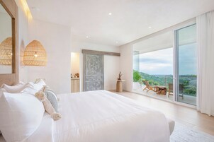 Um quarto espaçoso com uma cama grande, vista para o verde através de uma porta de vidro deslizante e uma varanda com área de estar externa.