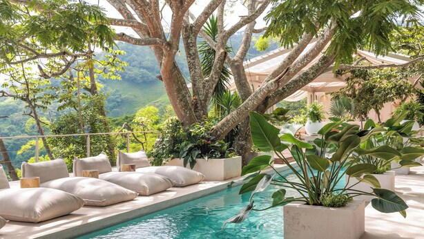 Zona de piscina con vistas a las montañas, rodeada de grandes macetas con plantas y asientos acolchados.