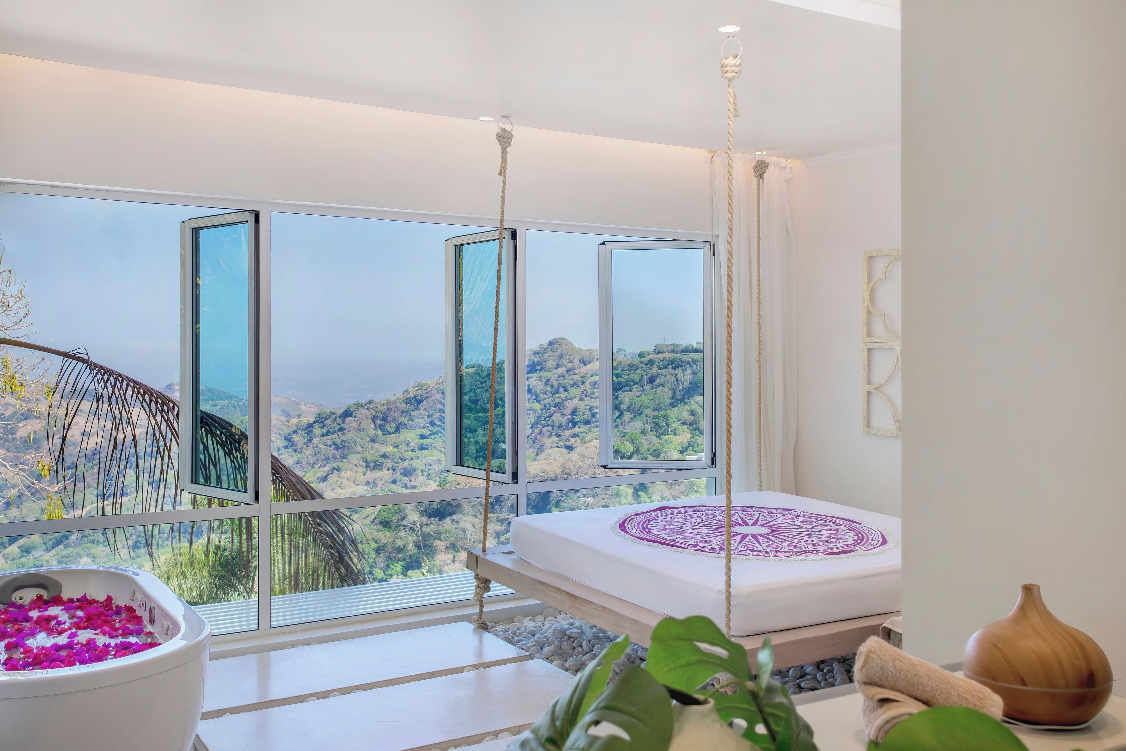A bedroom with a large window, a bed with a decorative headboard, a hanging chair, and a view of a mountainous landscape.