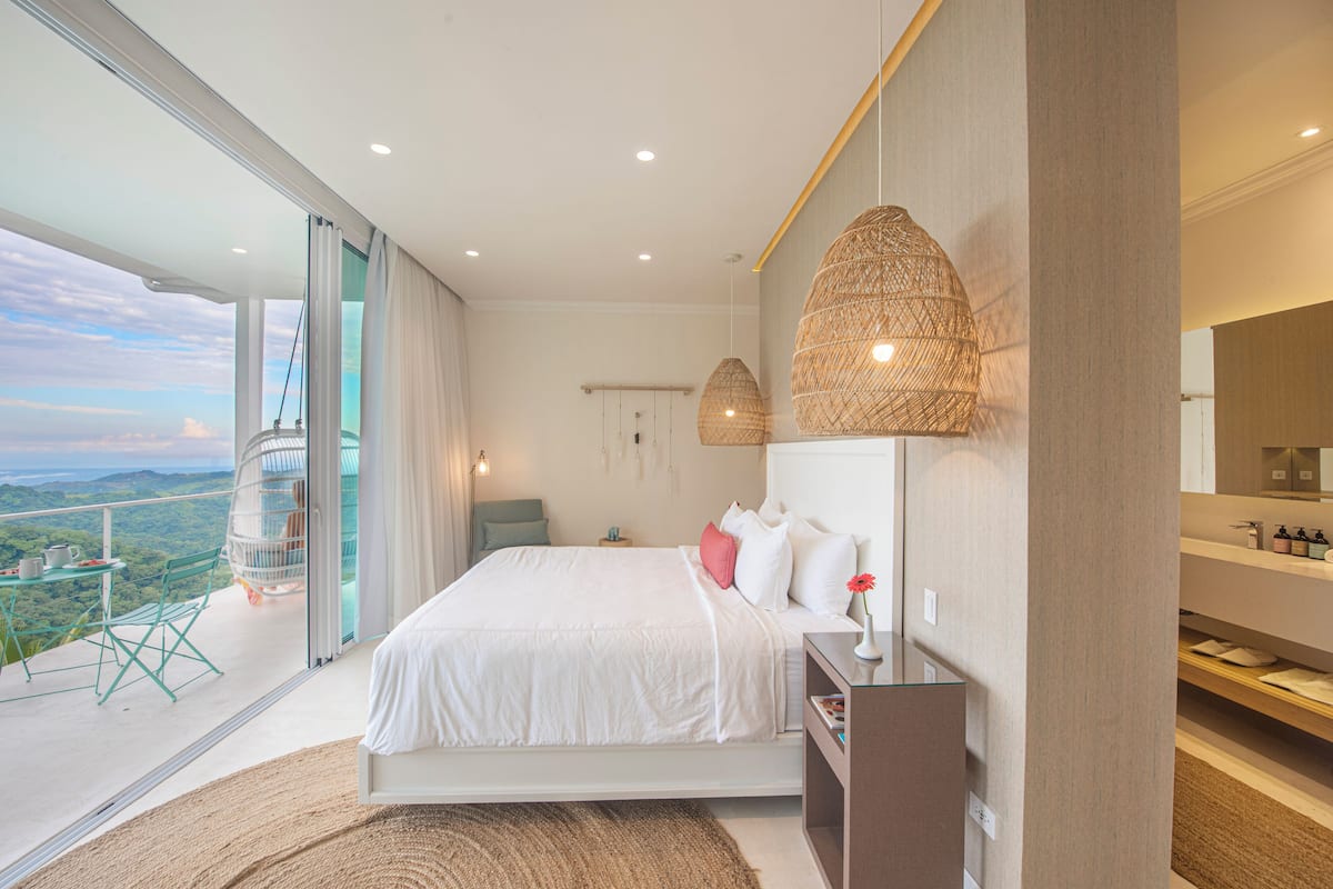 A modern bedroom with a large bed, a balcony with a view, and a bathroom with a mirror and shelves.