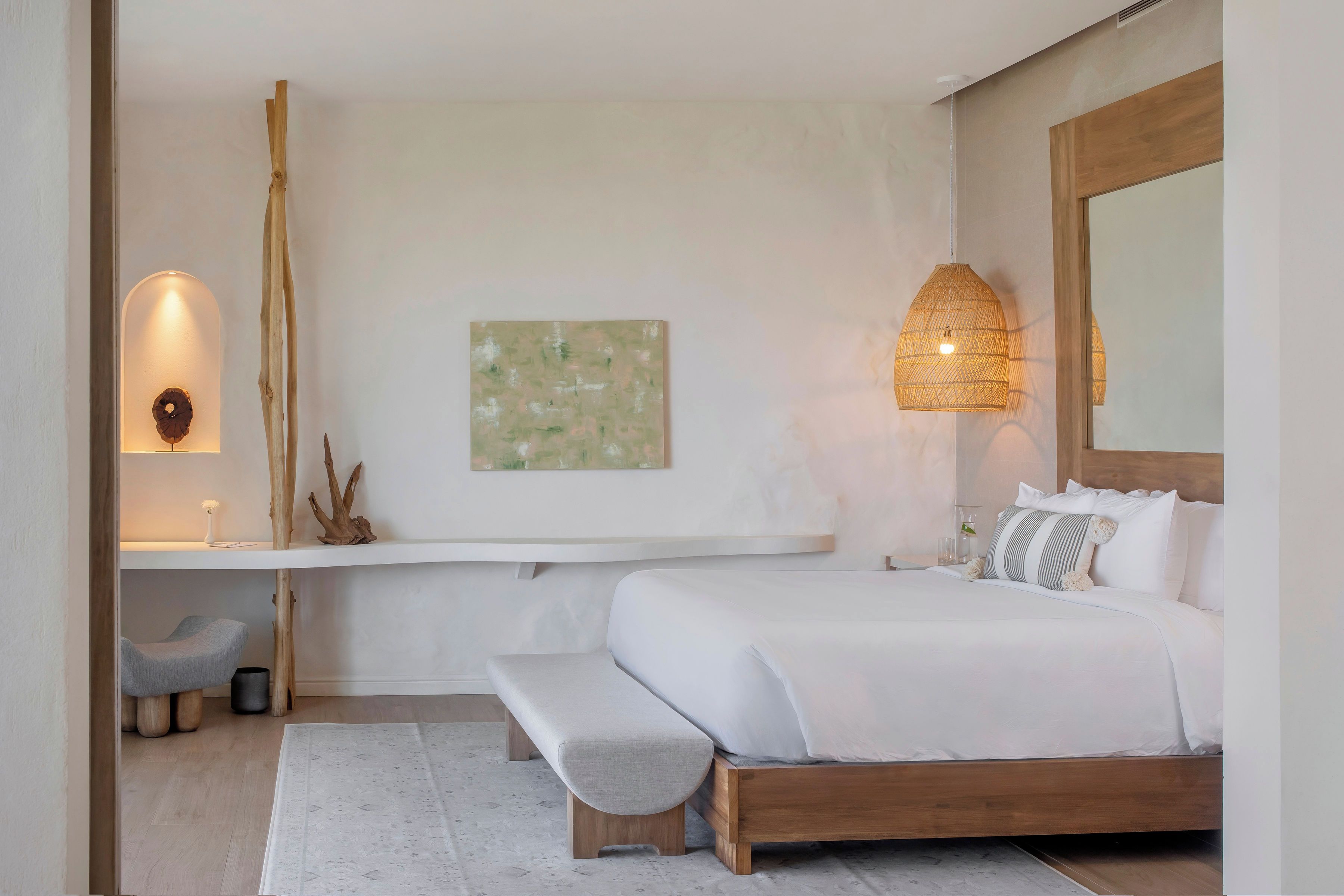 A bedroom with a wooden bed, a bench, a bedside table, a wall-mounted lamp, and a painting.