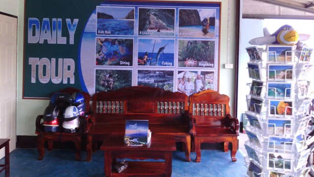 Una habitación con un letrero que dice 'DAILY TOUR', un cartel que reza 'HONESTY IS OUR POLICY' y una exposición de fotografías de viajes. Hay dos sofás de madera, una mesa con un folleto y una pila de postales.