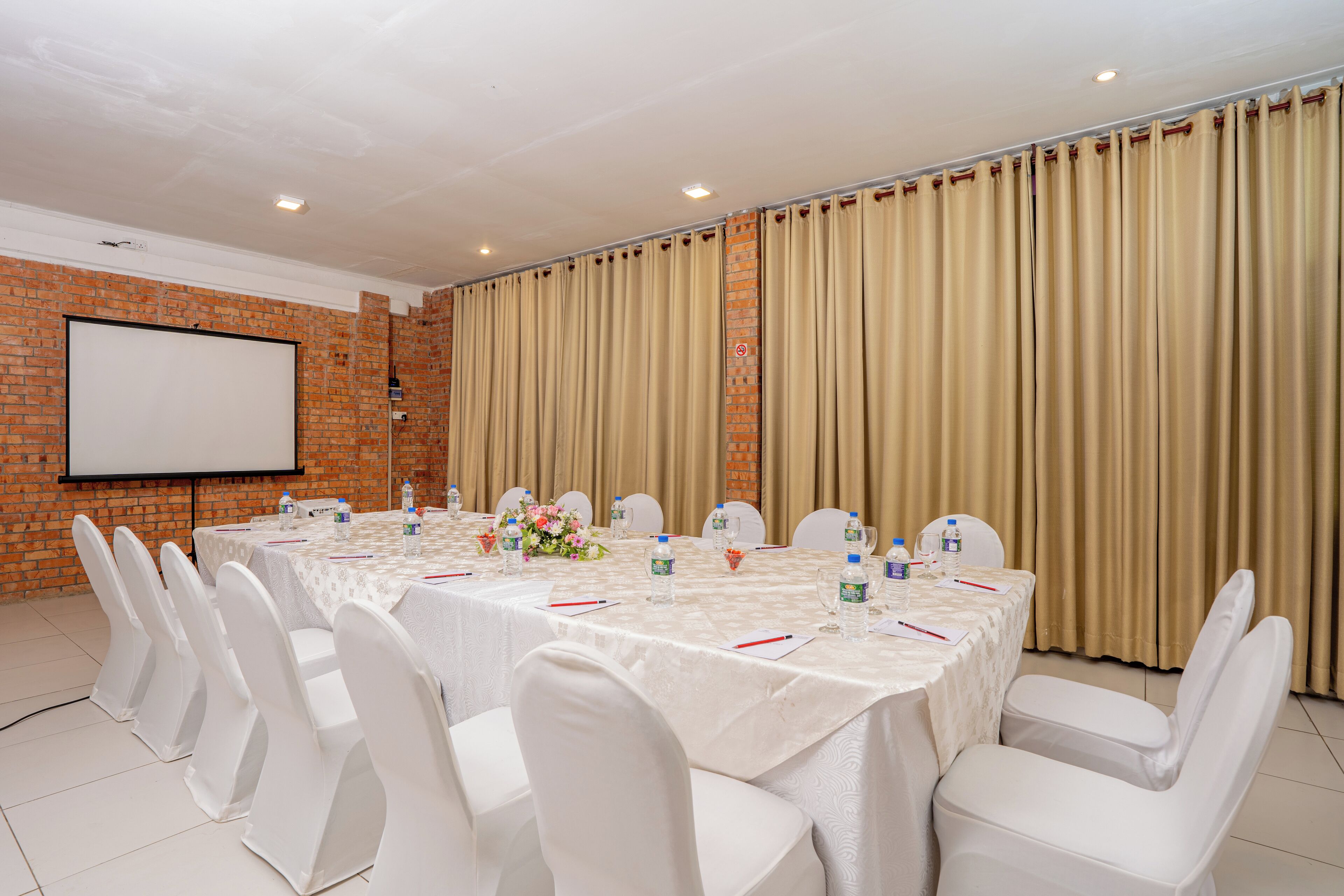A conference room with a long table, white chairs, a projector screen, and a brick wall.