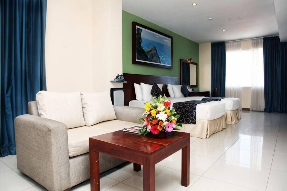 A hotel room with a sofa, a wooden coffee table with a flower arrangement, a bed with a headboard, and a painting on the wall.