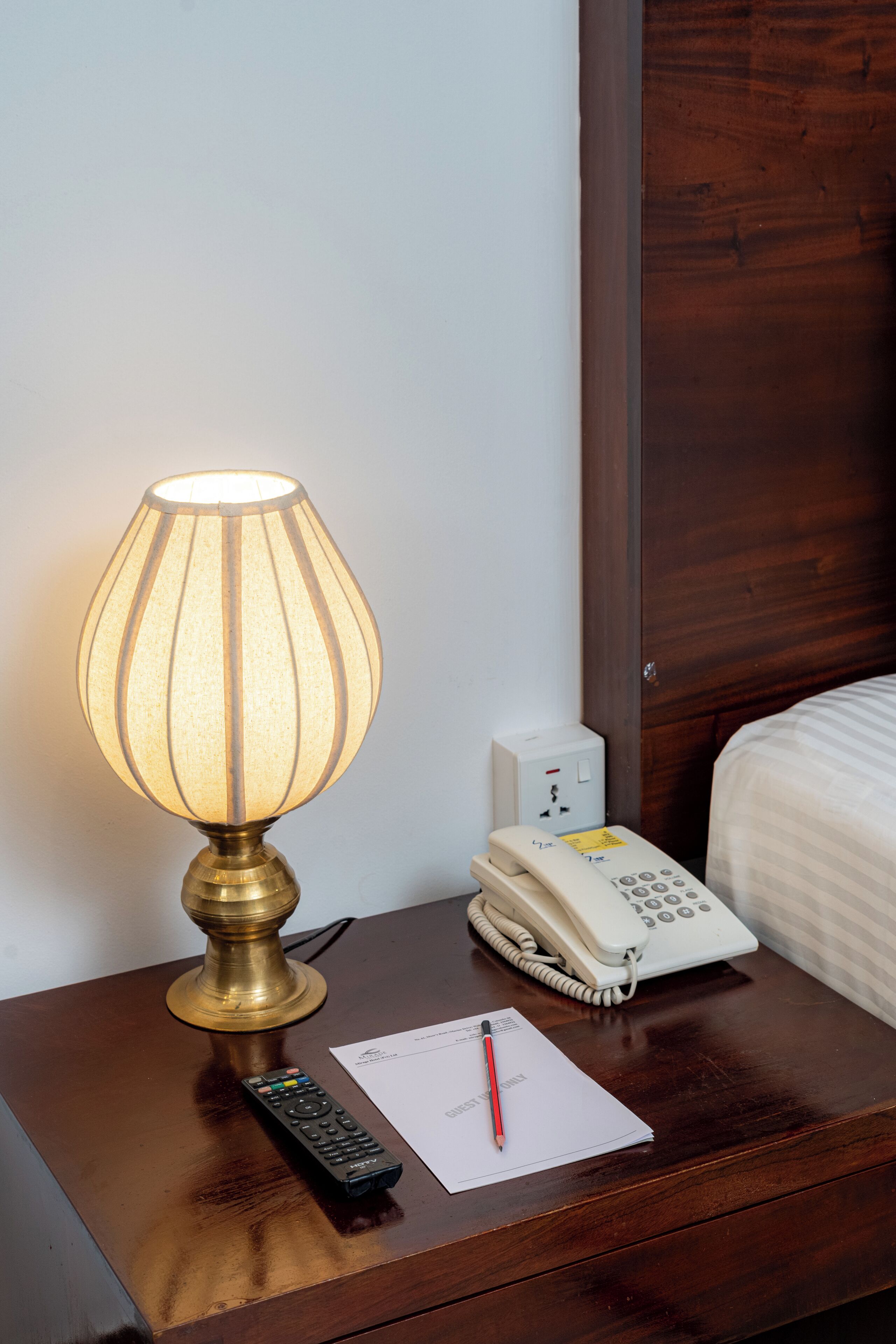 Une table de chevet avec une lampe, un téléphone, une télécommande et une feuille de papier sur laquelle se trouve un stylo.