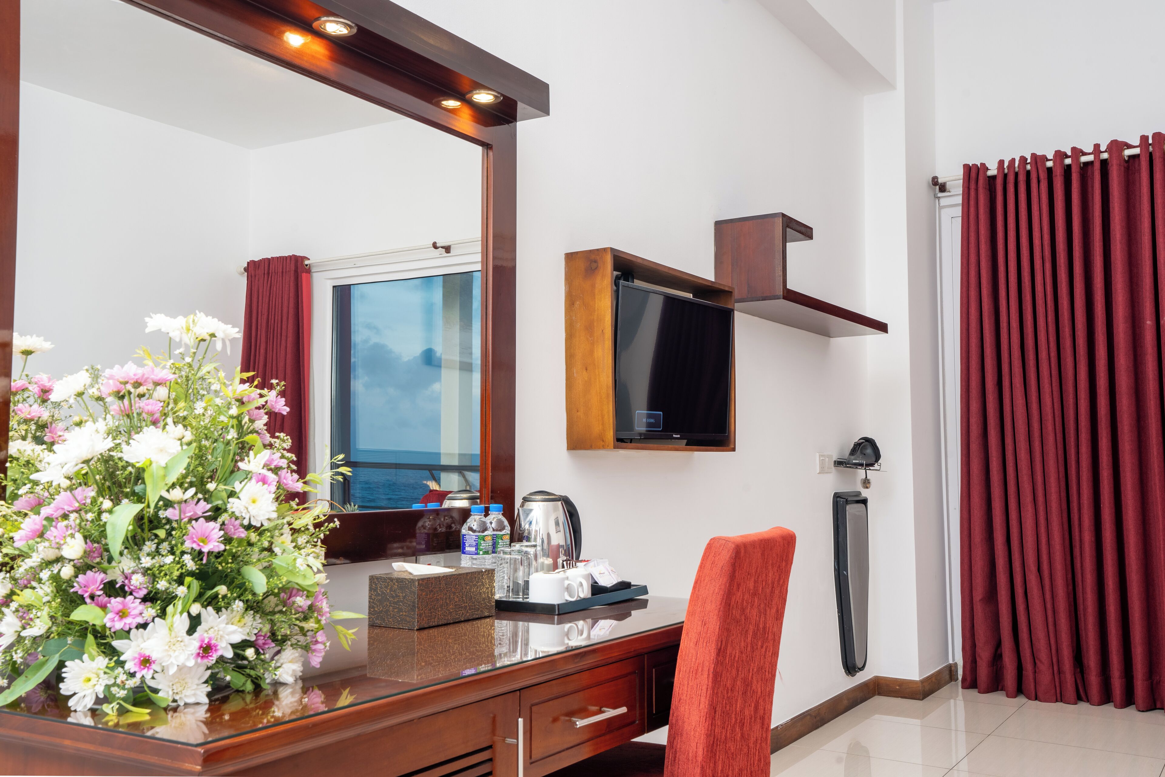 A hotel room with a wooden desk, a large mirror, a television, a red chair, a vase of flowers, and a view of the sea.