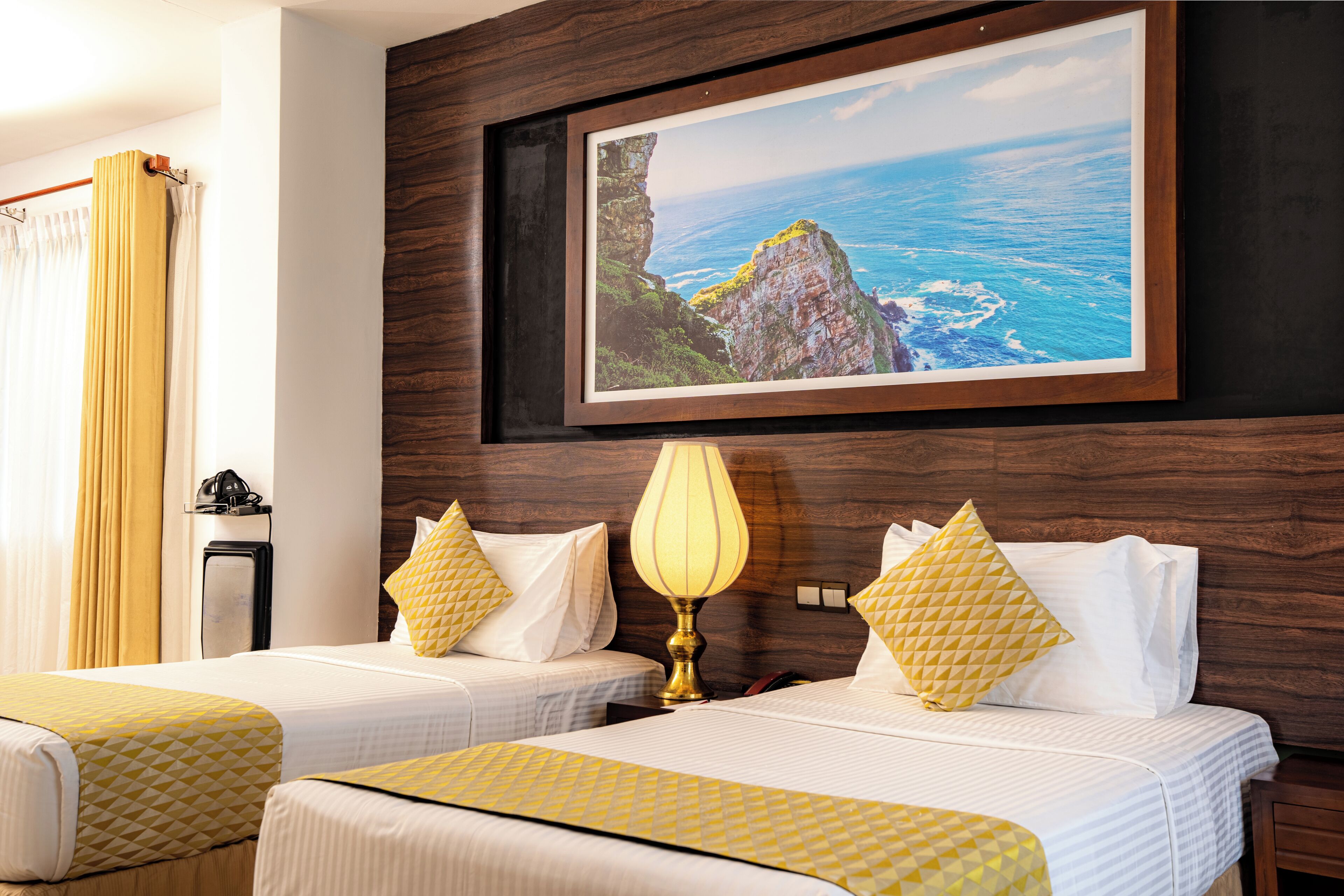 Ein Hotelzimmer mit zwei Betten, einem gerahmten Bild von einer Küstenlandschaft, einer Nachttischlampe und einem hölzernen Kopfteil.