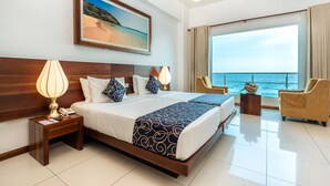 Habitación de hotel con una cama grande, dos mesitas de noche, una silla, un ventanal con vista al mar y un cuadro en la pared.