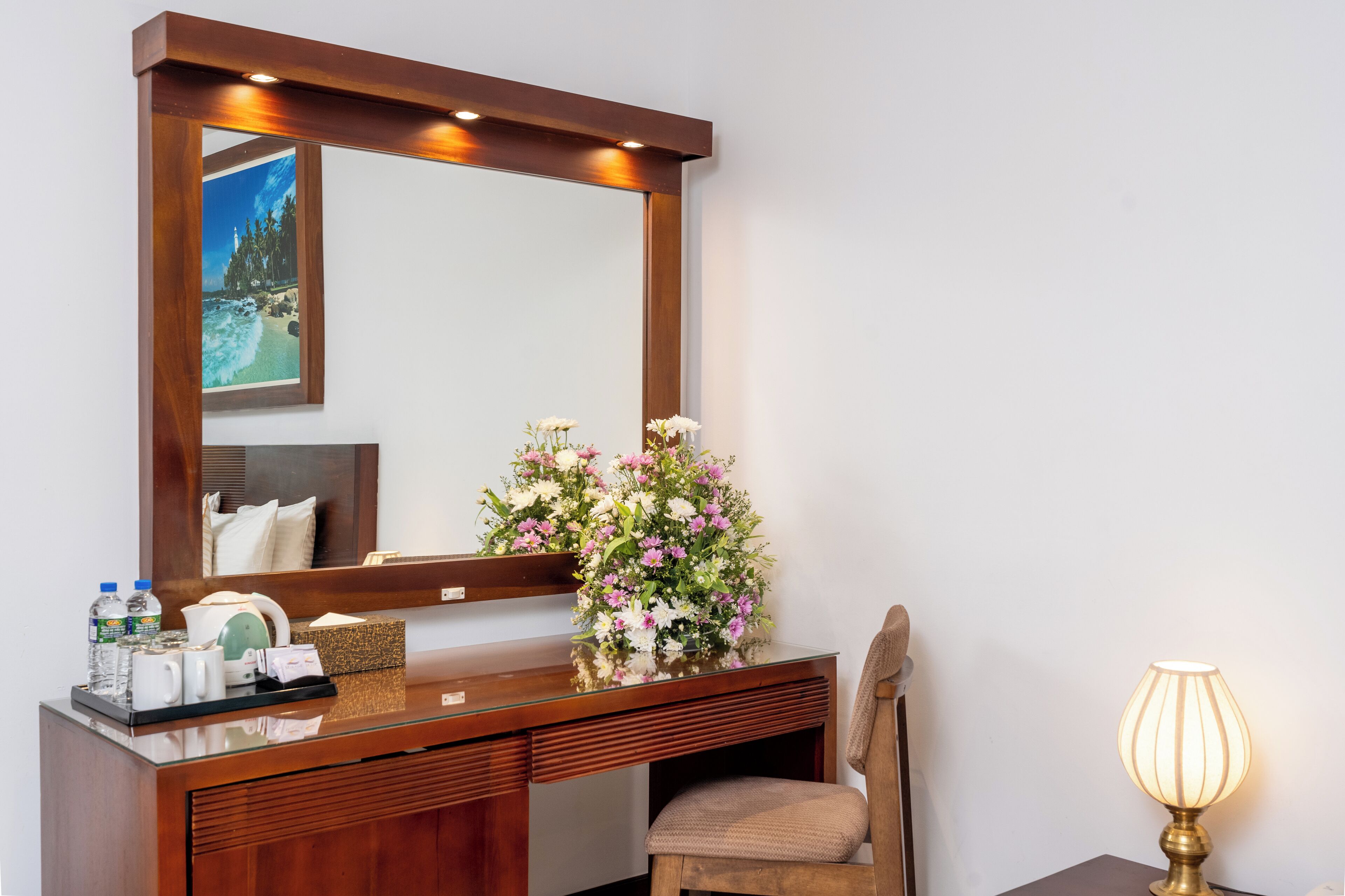 Quarto de hotel com escrivaninha de madeira, espelho, cadeira, luminária de cabeceira e um arranjo floral.