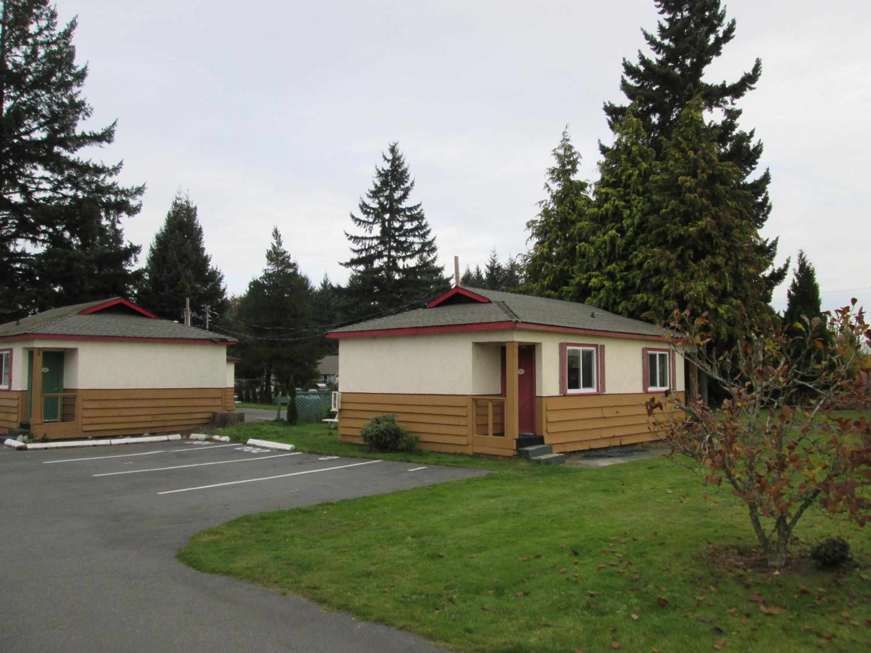 Two small cabins with a red roof, a parking lot, and a grassy area with trees.