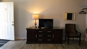 Quarto com um armário de madeira, televisão, luminária, cadeira, porta e espelho na parede.