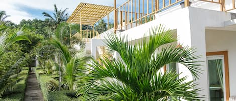 Un edificio moderno con fachada blanca, un balcón con barandillas de madera y abundante vegetación, incluyendo palmeras.