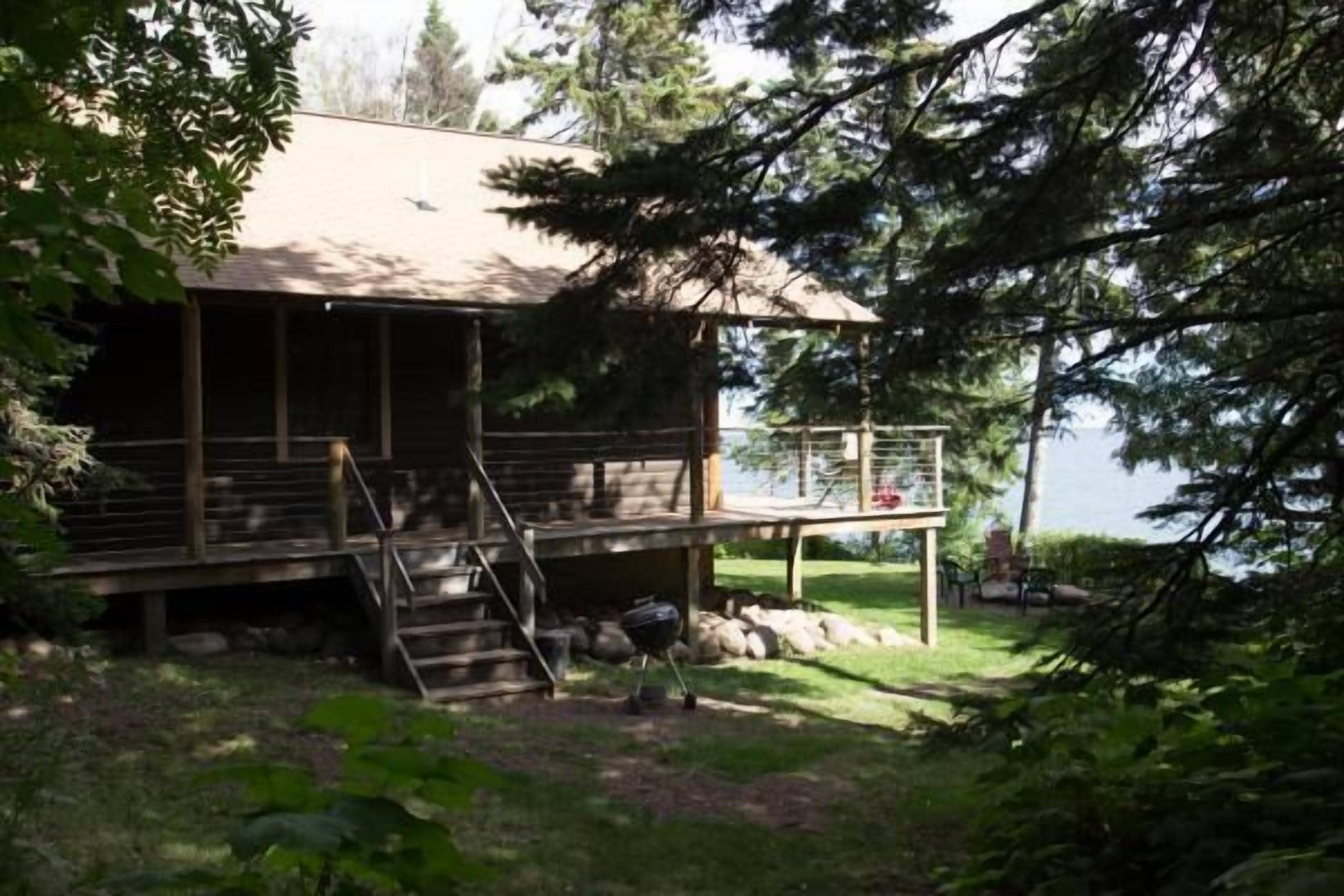 A cabin with a deck and stairs leading to a covered area, surrounded by trees and a lake view.