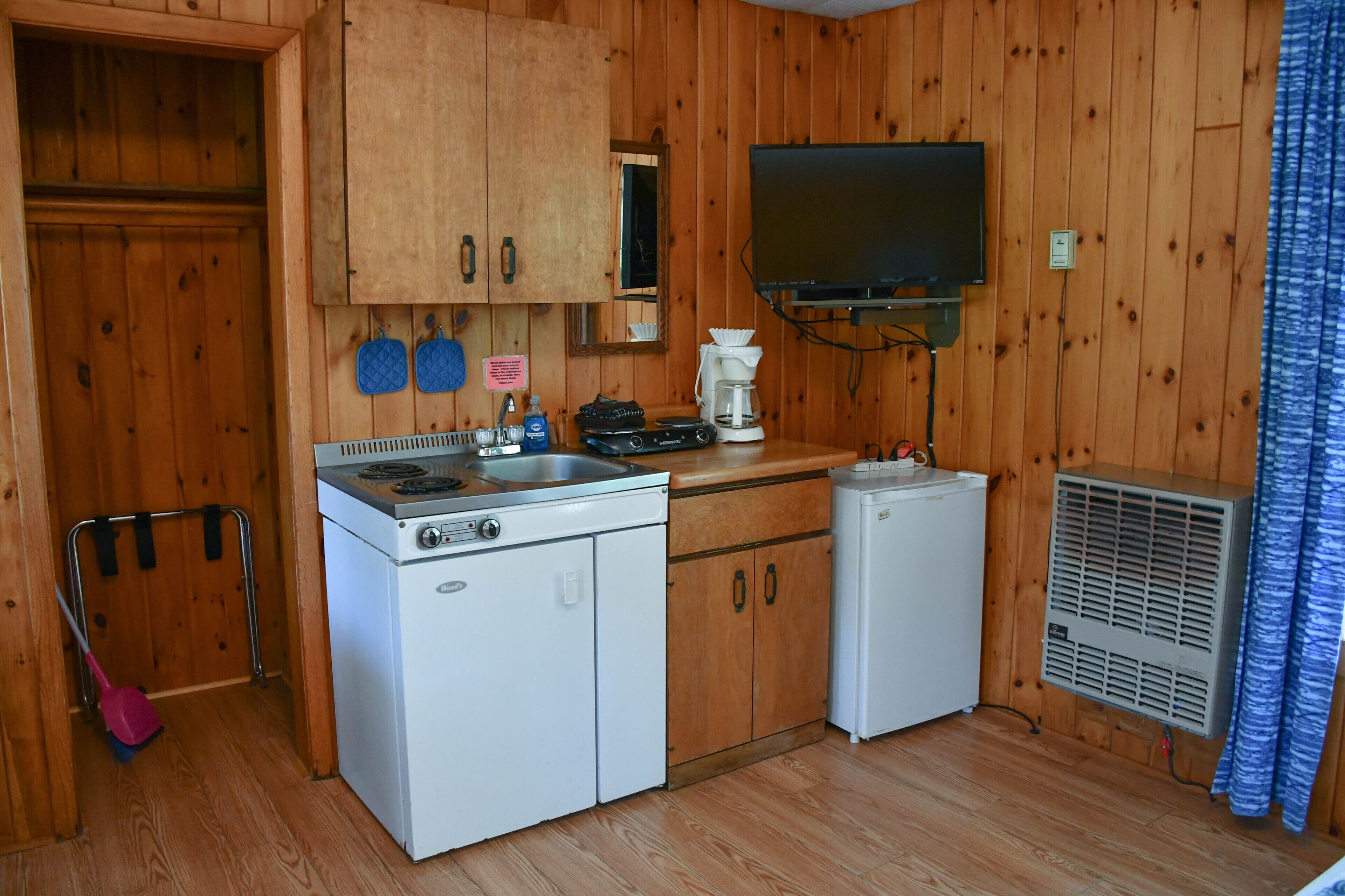 A compact kitchen with wooden walls, a stove, a refrigerator, a coffee maker, and a TV mounted above the counter.