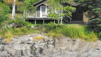 A cabin with a deck, surrounded by dense vegetation and a rocky shoreline.