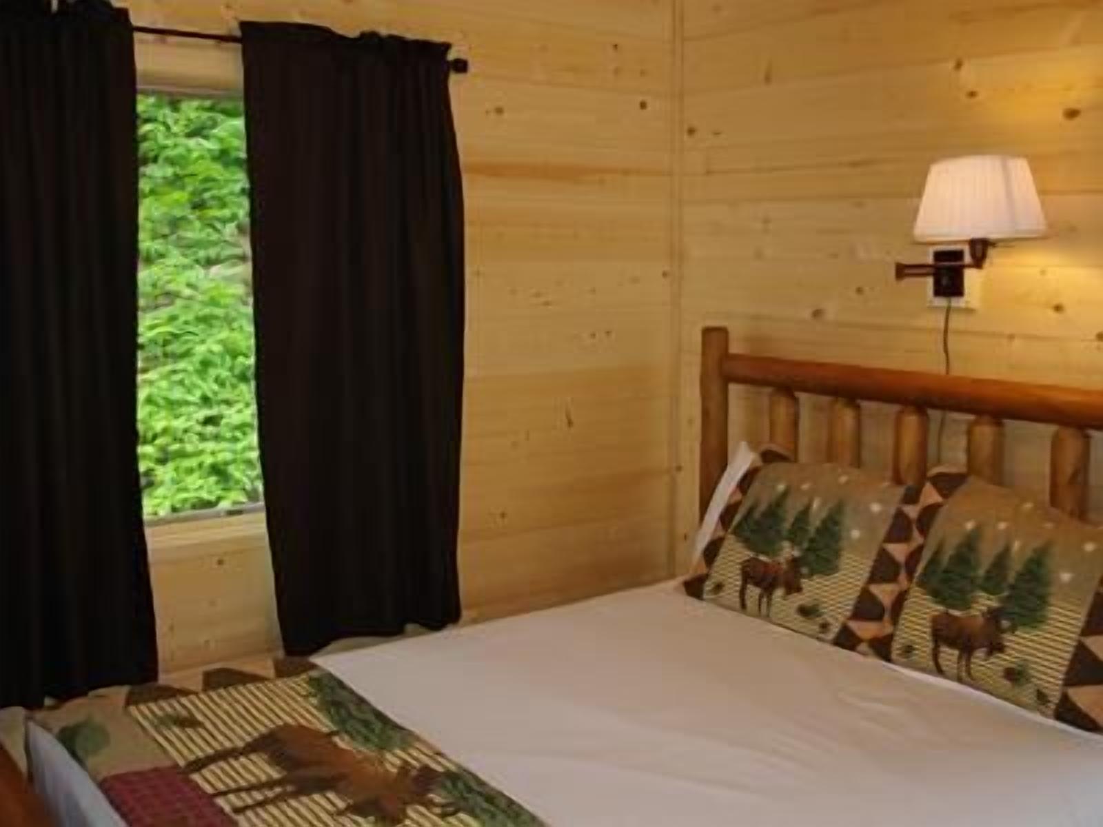 Un dormitorio de cabaña de madera con cama, cortinas, lámpara y ventana con vistas a la vegetación.