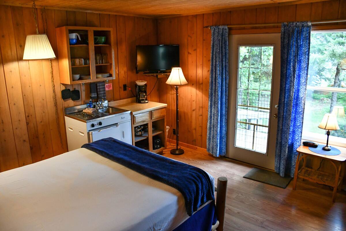 Une chambre dans un chalet en bois, comprenant un lit, un coin cuisine, une télévision et une porte donnant sur un balcon.